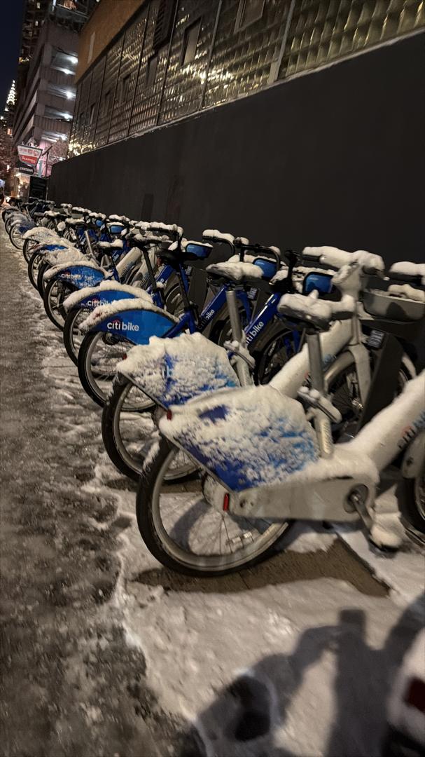 A row of Citi Bikes covered in fresh snow on an Astoria street at night